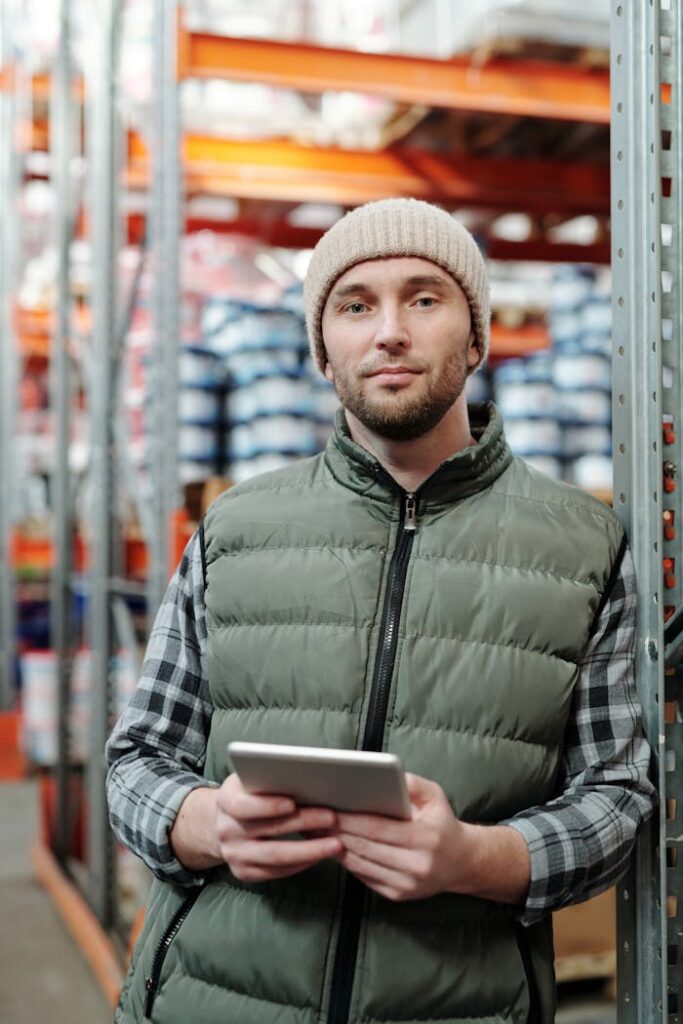 A man wearing a beanie and vest holding a tablet in a warehouse setting.